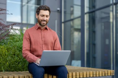 Olgun iş adamı dışarıda dizüstü bilgisayarla çalışır, dışarıda çalışma alanının tadını çıkarır. Günlük kıyafet, üretken ruh hali ve modern ortam, çağdaş iş yaşam tarzını yansıtıyor. Rahatlamış odak göstergesi.