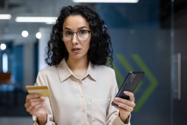 Ofisteki İspanyol iş kadını kredi kartı ve akıllı telefon tutuyor, şaşırmış görünüyor. Çevrimiçi işlemler konusunda kafası karışmış ya da endişeli görünüyor. Arkaplanda modern ofis alanı.
