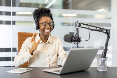 Kulaklıklı neşeli kadın, ofisteki podcast 'i mikrofon, dizüstü bilgisayar ve tabletle kaydediyor. İçerik oluşturucu, kusursuz sanal bağlantının keyfini çıkarıyor.