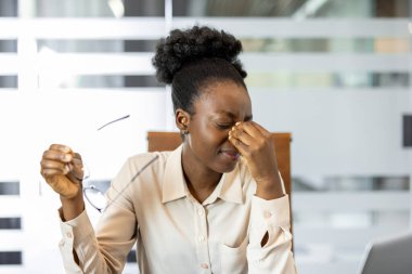 Modern ofis ortamında stres ve yorgunluk yaşayan iş kadını. Gözlerine masaj yaparken gözlük tutuyor, duygusal gerginlik ve iş yükü baskısı taşıyor..