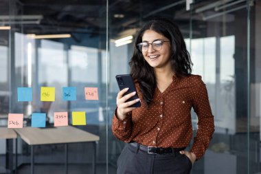 Modern ofiste akıllı telefona bakarken gülümseyen Latin Amerikalı kadın iş kadını. Çalışma ortamı, cam duvarda görünen yapışkan notlar, verimliliği ve yeniliği temsil ediyor.