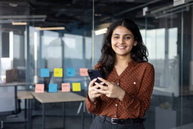Ofisteki Latin Amerikalı iş kadını elinde telefonla, güvenle gülümsüyor. Cam duvarlar, yapışkan notlar işbirliğini, modern iş yerini akla getiriyor. Teknolojinin ideal temaları, profesyonellik, mutluluk.