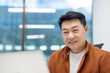 Asyalı iş adamı modern ofiste dizüstü bilgisayarla çalışırken kendine güvenerek gülümsüyor. Şirket ortamında mesleki başarı ve pozitif iş ortamı tasvir eder.