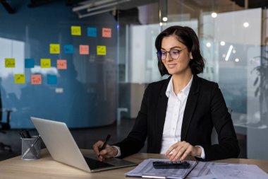 Modern ofiste çalışan iş kadını, dizüstü bilgisayara odaklanmış. Profesyonel ayarlar, duvarında yapışkan notlar, masasında belgeler. Ofis ortamı, yaratıcı planlama kavramı.