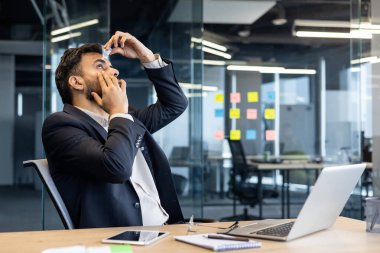 Takım elbiseli bir ofis çalışanı, masadaki göz damlalarını dizüstü bilgisayar ve telefonla, çoklu görev, stres atma ve modern işyerindeki dijital gerginliği simgeliyor..