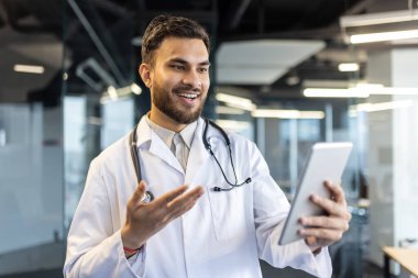 Kendine güvenen doktor modern ofiste tablet üzerinde sanal danışmanlık yapıyor. Gülümseyen, meşgul bir profesyonel teknolojiyi uzaktan sağlık hizmetleri için kullanıyor. Beyaz önlük, steteskop tıbbi titreşimleri arttırır..