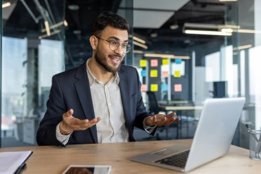 Modern ofisteki iş adamı dizüstü bilgisayarla sanal toplantı yapıyor. Kendine güvenen ifade, açık jestler iletişimi vurgulamak, profesyonelliği vurgulamak. Masadaki akıllı telefon dijital çalışma alanını gösterir.