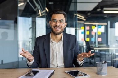 Modern ofisteki kendine güveni tam bir işadamı, içten gülümseyen, sohbete dalan. Masada bulunan telefon ve tablet gibi teknoloji, profesyonel çevreyi, iletişimi ve açıklığı yansıtıyor.