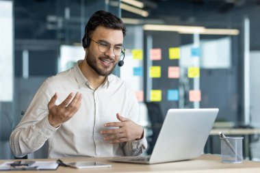 Sanal toplantıda kulaklık takan bir iş adamı bilgisayarla uzaktan sohbete girişiyor. Modern ofis kurulumu dijital iletişim verimliliğini vurguluyor. İş bağlamına uygun.