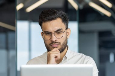 Gözlüklü bir adam ofis ortamında bilgisayar ekranına odaklanıyor. Ciddi ifade derin düşünce ya da analiz önerir. Modern çalışma ortamı. Anahtar sözcükler: ofis, odaklanma, profesyonel.