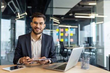 Akıllı ofisteki kendine güvenen genç iş adamı dizüstü bilgisayarı, kalemi ve masasında defteri var. Kameraya gülümsüyor, verimliliği, başarıyı, profesyonel kıyafeti temsil ediyor. Modern iş ortamı.