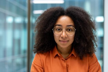 Kıvırcık saçlı, kendine güvenen bir kadın parlak ofiste duruyor. İş ortamlarında profesyonellik, güçlendirme ve çeşitlilik temaları için ideal.