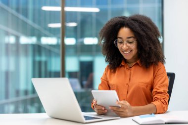 Modern ofiste dijital tablet ve laptop kullanan gülümseyen profesyonel bir kadın. İş hayatı dengesi ve verimliliği vurgulayarak, teknoloji ve iletişimle meşgul olmak.