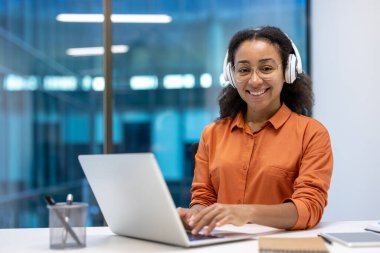 Laptop kullanan, kulaklık takan genç bir kadın. Profesyonel, işine odaklanmış, güvenle gülümseyen. Çalışmak, üretkenlik ve modern çalışma alanı sergilemek..