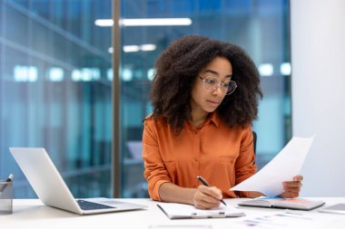 Masada belgelerle çalışan profesyonel bir kadın. Bilgileri laptop ve kağıtlarla gözden geçirir. Bu ayar modern ofiste iş, konsantrasyon ve verimliliği aktarır..