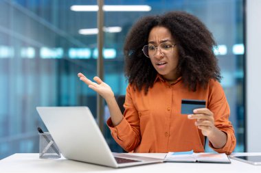 Ofiste dizüstü bilgisayar kullanırken kredi kartı tutan kafası karışmış bir kadın. Hüsrana uğramış ifade, çevrimiçi alışveriş veya ödeme sorunlarıyla ilgili sorun yaratarak endişe ve tatminsizlik yaratıyor.