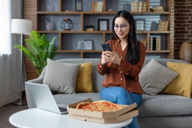 Asya 'lı bir kadın oturma odasında akıllı telefon kullanırken pizzadan zevk alıyor. Masada dizüstü bilgisayarla rahat bir atmosfer, ev ortamında birden fazla görev, konfor ve teknoloji kullanımı gösteriyor.