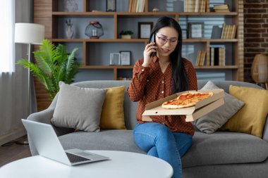 Asyalı bir kadın evde pizza kutusu tutarken telefonda konuşmaktan zevk alıyor, rahat bir atmosfer yaratıyor. Masadaki dizüstü bilgisayar uzaktan kumandalı olduğunu gösteriyor. Gelişigüzel ama verimli bir ev ortamı..