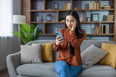 Evde akıllı telefon kullanan Asyalı bir kadın. Kanepede oturmak, telefonda beklenmedik mesajlar okumak, güvensizlik içinde yüz yüze. Ev dekoru ile günlük ayarlar.