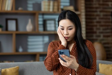 Evdeki Asyalı kadın elinde akıllı telefon tutarken şaşırmış görünüyor. Modern yaşam tarzı, ev içi ve iletişim konsepti. Rahatlamış ama endişeli ifade.