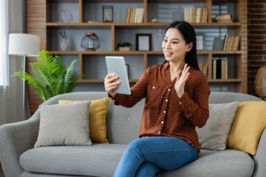 Asyalı kadın gülümsüyor, kanepeye oturmuş sanal konuşma için tablet kullanıyor. Selamlaşarak el sallamak, evde rahatlığın tadını çıkarmak. Sanal iletişim, teknoloji, bağlantı vurgulanıyor.