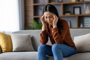 Asyalı kadın baş ağrısı çekiyor, evdeki kanepede oturuyor. Parmaklarıyla tapınaklara baskı yapıyor, stresli ve rahatsız görünüyor. Günlük kıyafetler, iç mekan dekorları, gerginlik ve yorgunluk..