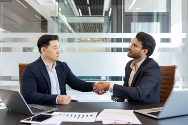 Modern ofiste iş adamları el sıkışıyor. Resim ortaklığı, güveni, başarılı anlaşmayı yakalar. Masadaki dizüstü bilgisayarlar, çizelgeler, belgeler profesyonel çevreyi ön plana çıkarıyor. Takım çalışması kavramı, işbirliği