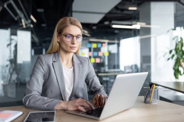 Modern ofiste dizüstü bilgisayar kullanan, işine odaklanmış olgun bir iş kadını. İş kıyafetleri profesyonelliği ve bağlılığı yansıtır. Uzmanlık ve üretkenlik gösteren görevlerle meşgul.