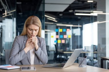 Ofisteki masada öksüren yetişkin bir iş kadını dizüstü bilgisayarı ve telefonu yanında. Pek iyi görünmüyor, mesleki çevrede rahatsızlığını ve endişelerini dile getiriyor..
