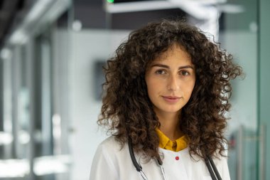 Hastane ortamında kıvırcık saçlı, steteskop takan profesyonel bir kadın doktor. Sağlık, güven ve sağlık hizmetlerini temsil ediyor. Tıbbi ve mesleki temalar için ideal görüntü.