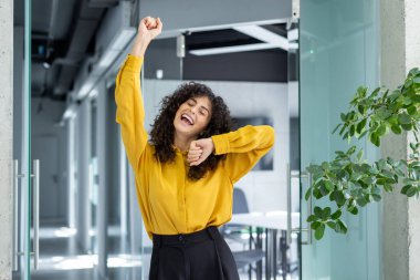 Happy woman celebrating success in modern office. Expressive joy, raised arm, signifying accomplishment and positive energy. Lively atmosphere with indoor plants and natural light. clipart