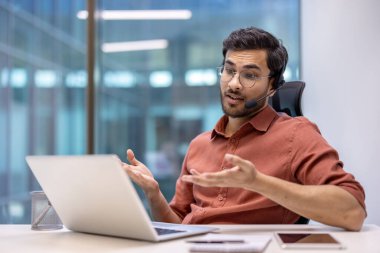 Kulaklık takan genç adam görüntülü arama yapıyor, dizüstü bilgisayar kullanıyor, el kol hareketi yapıyor. Modern ofis ortamında oturan, masadaki dijital cihazlar, iletişim ve teknoloji kavramlarının iletimi.