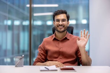 Ofisteki işadamı kameraya el sallıyor, kendine güvenini ve dostluğunu gösteriyor. Not defteri ve akıllı telefonuyla masada oturuyor, şık günlük kıyafetler giyiyor. Pozitif iş yeri iletişimi.