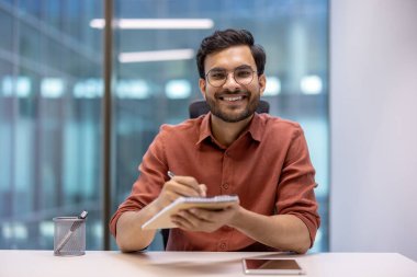 Ofiste oturan gülümseyen adam, elinde not defteri, toplantı sırasında not almaya hazır. Masadaki günlük gömlek, gözlük ve tablet modern profesyonel ortamı gösteriyor. Kendinden emin ve motive..