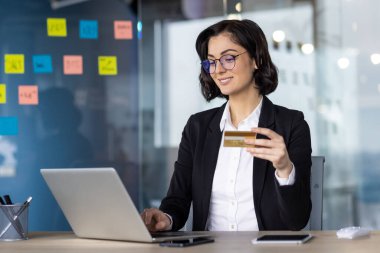 İş kadını online alışveriş için dizüstü bilgisayar kullanıyor, kredi kartı tutuyor, kendine güvenerek gülümsüyor. Laptop, telefon gibi dijital aygıtlarla modern ofis ayarları.