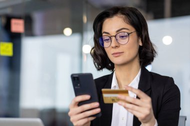 Ofisteki kadın akıllı telefon ve kredi kartıyla online ödeme yapıyor. Gözlüklü ve iş kıyafetli profesyonel görünüm, iş yerindeki faaliyetlere ve güvene işaret ediyor.