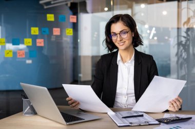Ofisteki iş kadını belgeleri inceliyor, güven ve profesyonelliği gösteriyor. Dizüstü bilgisayarla masa başında çalışıyor, iş yerinde liderlik gösterisi yapıyor. İş veya ofis temaları için ideal.