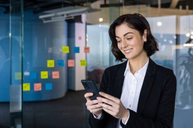 Ofisteki profesyonel iş kadını iletişim için akıllı telefon kullanıyor. Resmi iş kıyafetleri içinde, telefonla uğraşırken gülümsüyor, üretkenliğini ve bağlantısını gösteriyor..