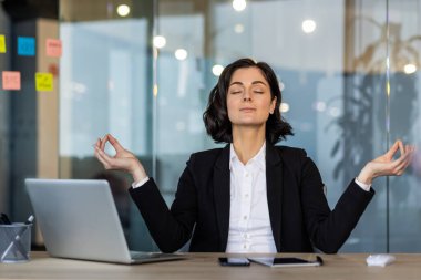 İş elbiseleri giymiş bir kadın ofis masasında meditasyon yapıyor, gözleri kapalı, sükuneti ve farkındalığı kucaklıyor. Yakınlardaki dizüstü bilgisayar ve telefon, iş ve rahatlama arasındaki dengeyi vurguladı.