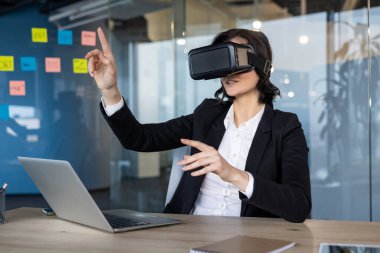 Sanal gerçeklik kulaklığı takan iş kadını interaktif dijital sunumla meşgul oluyor. VR teknolojisinden yararlanarak, bir ofis masasında işyeri çözümlerini araştırıyor..