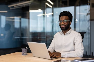 Modern ofis ortamında dizüstü bilgisayarda daktilo kullanan Afro-Amerikalı bir iş adamı. Gözlüklü, beyaz gömlekli, kendinden emin ve profesyonel görünüyor. Çevre cam duvarlara yansıyor, parlak bir hava yaratıyor..