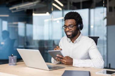 Afrikalı Amerikalı iş adamı dizüstü bilgisayar ve not defteri kullanarak çevrimiçi öğrenme işine girdi. Kulaklık takarak, modern ofis ortamında odaklanmış ve gülümsüyor..