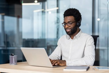 İş yerinde oturan Afro-Amerikalı adam iletişim için dizüstü bilgisayar ve kulaklık kullanıyordu. Odaklanma, teknoloji, uzaktan çalışma verimliliği. Modern iş kavramları ve profesyonel ortamlar için ideal.