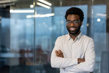 Modern ofiste kollarını kavuşturmuş gözlük ve gömlek takan Afro-Amerikalı bir işadamı. Kendine güvenen ifade, iş ortamında profesyonelliği ve başarıyı yansıtır, pozitif enerji yayar..