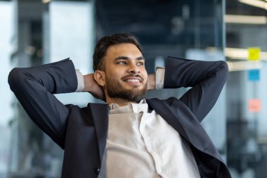 Modern bir ofis ortamında bir işadamı ellerini başının arkasına koyup rahatça oturur. Rahatlamış tavrı ve gülüşü başarılı bir işi yansıtan güven ve memnuniyeti ifade ediyor.