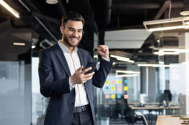 Takım elbiseli bir işadamı, elinde akıllı bir telefonla modern bir ofiste heyecan ve kutlama sergiliyor. Onun mutlu ifadesi bir iş ortamında başarı ve başarıyı vurguluyor..