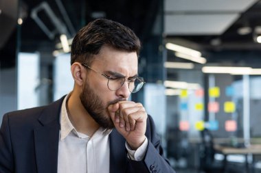 Bir işadamının ofis ortamında öksürdüğü, soğuk algınlığı veya grip belirtileri gösterdiği görülüyor. Gözlük ve takım elbise takmış, elini ağzına dayayıp hastalığını ve rahatsızlığını gösteriyor..