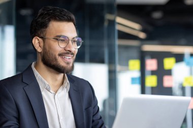 Kendine güvenen bir işadamı, parlak bir ofis ortamında dizüstü bilgisayarıyla çalışırken gülümsüyor. İfadesi profesyonelliği yansıtıyor ve modern ve üretken bir kurumsal çevreye odaklanıyor..