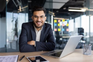 İş adamı bir şirket ofisindeki masasında güven içinde oturuyor. Gülümsüyor, takım elbise giyiyor, önünde dizüstü bilgisayar ve akıllı telefon ile profesyonelliği ve başarıyı yansıtıyor..