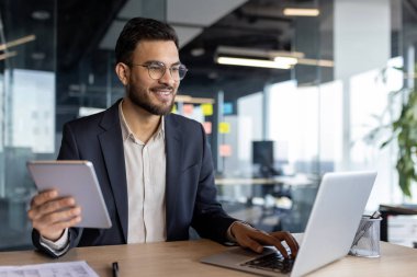 Bir işadamı, bir dizüstü bilgisayar ve tablet kullanarak modern bir ofiste oturuyor, üretkenliği ve dijital cihazlarla birden fazla görevi somutlaştırıyor. Kendine güveni ve dostça tavırları var.